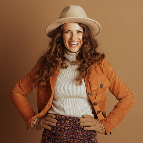 Woman wearing a beige hat, orange jacket, and patterned skirt against a brown background